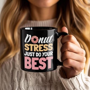 May include: Black ceramic mug with the words "Donut Stress Just Do Your Best" in white, tan, and pink. The word "Donut" has a graphic of a donut. The mug has a black handle and is held by a person.
