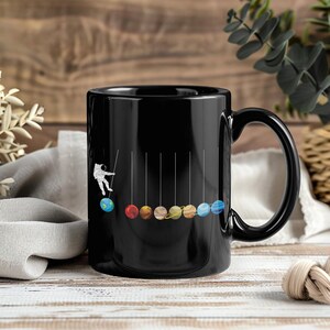 Könnte beinhalten: Schwarze Keramik-Tasse mit einem verspielten Design eines Astronauten und den Planeten des Sonnensystems. Die Planeten sind als farbige Kugeln dargestellt, die in einer Reihe hängen, wobei der Astronaut scheinbar mit ihnen interagiert. Eine lustige Geschenkidee zum Thema Weltraum.