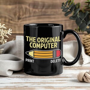 May include: Black ceramic mug featuring the text "THE ORIGINAL COMPUTER" in yellow. A pencil graphic is below the text, with "PRINT" and "DELETE" on either side. The mug has a black handle.