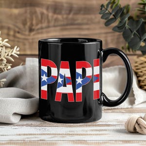 May include: Black ceramic mug with the word "PAPI" in large, bold letters. The letters are filled with the Puerto Rican flag design, featuring red stripes, white stars, and blue triangles. The mug has a black handle.