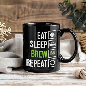 May include: Black ceramic mug with the text "EAT SLEEP BREW REPEAT" in white and green. The mug has icons of a plate, bed, hops, and a recycling symbol. The mug has a black handle. The background is a wooden surface.