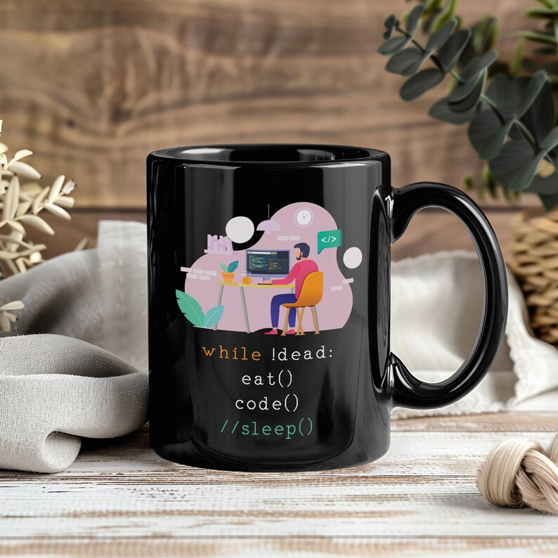 Puede incluir: Taza de cer&aacute;mica negra con un gr&aacute;fico de una persona codificando. La taza tiene el texto "while !dead: eat() code() //sleep()" en blanco. La taza tiene un asa negra.