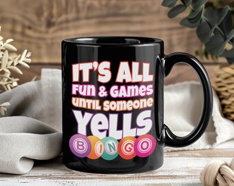 May include: Black ceramic mug with the text "IT'S ALL FUN & GAMES UNTIL SOMEONE YELLS BINGO" in white, pink, and purple. The word "BINGO" is spelled out with colourful bingo balls. The mug has a black handle.