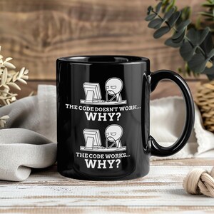以下が含まれることがあります： 白のグラフィックが描かれた黒いセラミックコーヒーマグ。コンピューターの前にいる人物と「THE CODE DOESN'T WORK... WHY?」と「THE CODE WORKS... WHY?」のテキストが描かれています。マグには大きなハンドルが付いています。
