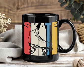 May include: Black ceramic mug featuring the word "SKATE" in red, blue, and yellow vertical stripes. A cream-colored roller skate is centered within the design. The mug has a black handle and is set against a wooden background.