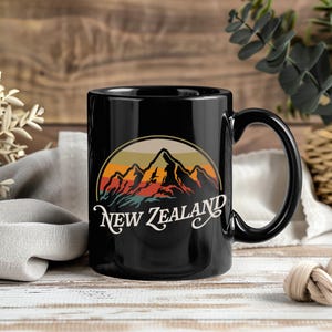 Op de afbeelding: Zwarte keramische mok met een berglandschapontwerp en de tekst "New Zealand" in witte letters. Het ontwerp heeft een zonsondergangkleurenschema van oranje, geel en bruin. De mok heeft een gebogen handvat.