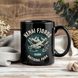 May include: Black ceramic mug featuring a circular design with the words "Kenai Fjords Alaska National Park." The design includes a mountain range, water, and orcas. The mug has a black handle and a glossy finish.