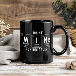 Puede incluir: Una taza de cerámica negra con texto blanco que dice I DRINK WINE PERIODICALLY. El diseño incluye tres elementos de la tabla periódica: W para Tungsteno, I para Yodo y Ne para Neón, junto con sus números atómicos y pesos atómicos.