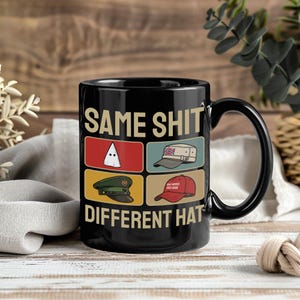 Op de afbeelding: Zwarte keramische mok met de tekst "SAME SHIT DIFFERENT HAT" en vier hoedillustraties. De hoeden zijn onder meer een witte puntmuts, een militaire pet, een rode baseballpet en een Britse militaire pet.
