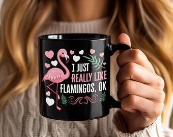 May include: Black ceramic mug featuring a pink flamingo illustration with the text "I JUST REALLY LIKE FLAMINGOS, OK". The design includes pink hearts, white hearts, and green leaves. The mug has a black handle.