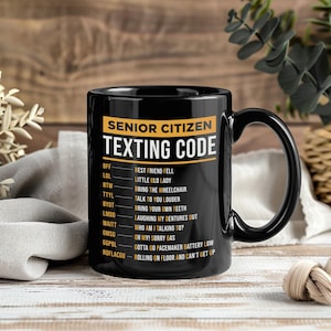 May include: Black ceramic mug with a large handle. The mug features the text "SENIOR CITIZEN TEXTING CODE" in white and yellow, with a list of texting acronyms and their meanings.