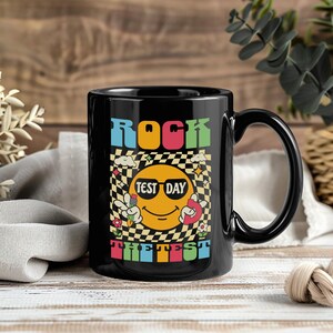 May include: Black ceramic mug with the words "ROCK THE TEST" in colorful block letters. The mug features a smiling emoji wearing sunglasses, holding a pencil and a peace sign, set against a checkered background.