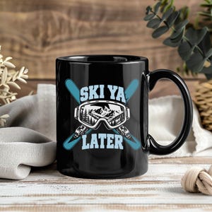 May include: A black ceramic mug featuring a graphic design of light blue crossed skis behind a pair of ski goggles. Inside the goggles is a black and white mountain landscape. The text "SKI YA LATER" is printed in light blue block letters.
