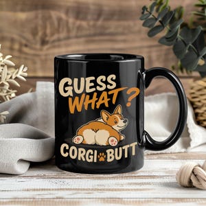 Puede incluir: Una taza de cerámica negra con una ilustración de dibujos animados de un perro Corgi acostado con la parte trasera hacia adelante. El texto impreso en naranja y blanco dice GUESS WHAT? CORGI BUTT, con una pequeña huella de pata naranja reemplazando la I.