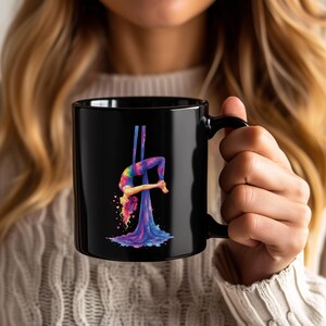May include: A black ceramic mug featuring a colourful graphic of an aerialist performing a backbend on blue and purple silk ribbons. The figure is depicted in vibrant shades of pink, yellow, and blue, with small heart shapes floating around the hair.