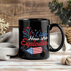 May include: Black ceramic mug with a patriotic design. The mug features the text "I like How she Explodes" in red, white, and blue, along with a firework illustration. The mug has a black handle and a glossy finish.