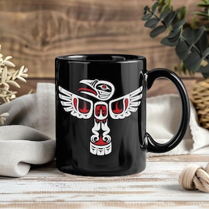 Puede incluir: Taza de cerámica negra con un diseño de tótem nativo americano blanco y rojo. El diseño incluye un pájaro con las alas extendidas y otras formas simbólicas. La taza tiene un asa negra y un acabado brillante, para bebidas calientes.