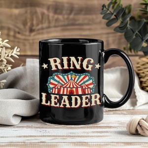 May include: A black ceramic mug featuring a graphic of a red, white, and blue circus tent. The text "RING LEADER" is printed in a bold, cream-colored serif font above and below the tent illustration, accented by small stars on each side.
