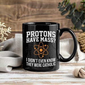 May include: A black ceramic mug featuring an orange atom graphic in the centre. The text printed in white reads: "PROTONS HAVE MASS? I DIDN'T EVEN KNOW THEY WERE CATHOLIC". The mug has a curved handle and a smooth, glossy finish on the exterior surface.