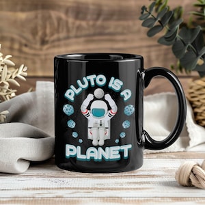 以下が含まれることがあります： 白とティール色の「PLUTO IS A PLANET」の文字が入った黒いセラミックマグ。宇宙飛行士のイラストと、いくつかの青い小惑星のグラフィックが特徴です。マグには黒いハンドルが付いています。