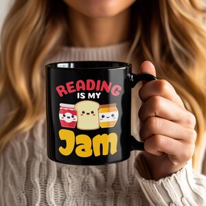 May include: Black ceramic mug with the text "Reading is my Jam" in red and yellow. The mug features illustrations of jam jars and a slice of toast. The mug is held by a person.