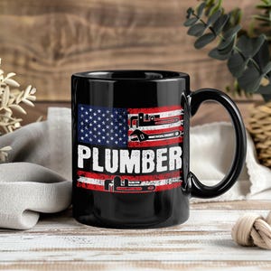 Könnte beinhalten: Schwarze Keramik-Tasse mit dem Wort "PLUMBER" in Weiß, einem amerikanischen Flaggen-Design und Schraubenschlüssel-Grafiken. Die Tasse hat einen schwarzen Henkel. Holz-Hintergrund.