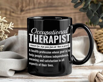 May include: Black ceramic mug with the text "Occupational THERAPIST" in white. The mug includes a definition of the profession. The mug has a black handle and is set against a wooden background.