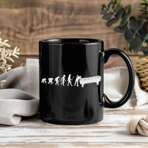 Può includere: Tazza da caffè nera con una grafica bianca che raffigura l'evoluzione di una persona che gioca a biliardo. La grafica mostra una progressione da figure simili a scimmie a una persona con una stecca da biliardo. La tazza ha un manico grande.