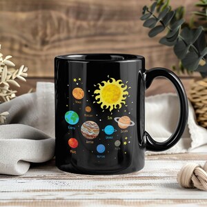 Peut inclure: Mug en céramique noire avec une illustration colorée du système solaire, comprenant le soleil et des planètes comme la Terre, Mars et Jupiter. Les planètes sont étiquetées. La tasse a une anse noire et une finition brillante.