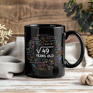 May include: Black ceramic mug with a large handle. The mug features colorful mathematical equations and the text "√49 YEARS OLD" in white. The design is a whimsical celebration of a milestone birthday.