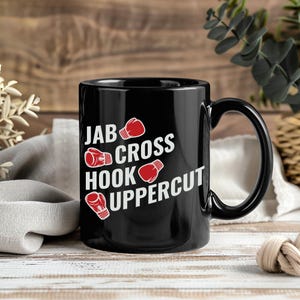 May include: Black ceramic mug with the words "JAB CROSS HOOK UPPERCUT" in white, along with red boxing glove illustrations. The mug has a black handle and is set against a wooden background.