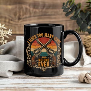 May include: A black ceramic mug featuring a graphic of two crossed revolvers over a retro-style sunset circle. The text reads I HAVE TOO MANY GUNS SAID NO ONE EVER in bold orange and yellow lettering, surrounded by decorative gold-colored scrollwork.