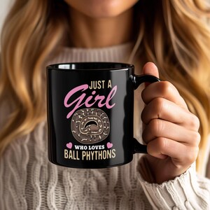 May include: Black ceramic mug with the text "Just a Girl Who Loves Ball Pythons" in pink and white. The mug features a graphic of a ball python. The mug is held by a person.