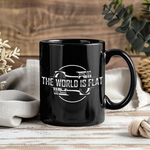 May include: A black ceramic mug with a white graphic of an engine piston and the text "THE WORLD IS FLAT". The mug has a large handle and is set against a wooden background.