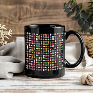 May include: Black ceramic mug featuring a collage of colorful, circular flag designs. The flags represent various countries and territories around the world. The mug has a large handle and is set against a wooden background.