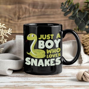 May include: Black ceramic mug featuring a cartoon snake and the text "JUST A BOY WHO LOVES SNAKES" in white and green. The mug has a black handle and is set against a wooden background.