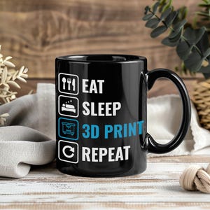Könnte beinhalten: Schwarze Keramik-Tasse mit den Worten "EAT SLEEP 3D PRINT REPEAT" in Weiß und Blau. Die Tasse hat einen schwarzen Henkel und steht vor einem Holzgrund.
