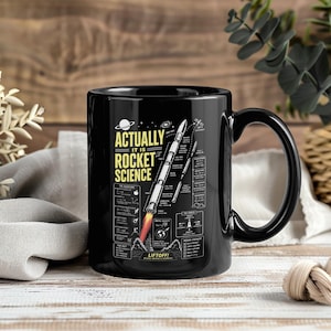 Może przedstawiać: Czarny ceramiczny kubek z biało-żółtą grafiką startującej rakiety, napisem "ACTUALLY IT IS ROCKET SCIENCE" i "LIFTOFF!" oraz różnymi schematami naukowymi, równaniami i listą kontrolną misji nadrukowanymi na przedniej stronie.