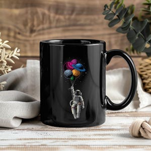 Peut inclure: Mug en céramique noire avec une illustration colorée d'un astronaute tenant des ballons en forme de planètes. Les ballons sont de couleurs violet, rose, bleu, jaune et vert. La tasse a une grande anse et est posée sur un fond en bois.