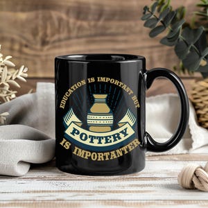 May include: Black ceramic mug with a curved handle. The mug features a design with the text "Education is important but Pottery is importanter" in a banner with a pottery wheel illustration in the center. The design is in shades of blue, tan, and white.