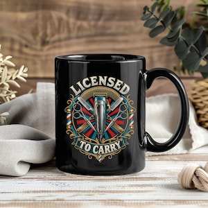 May include: A black ceramic mug featuring a graphic design with the text LICENSED TO CARRY. The illustration includes a central hair clipper, crossed scissors, and a comb, surrounded by a decorative circular frame with red and blue accents.