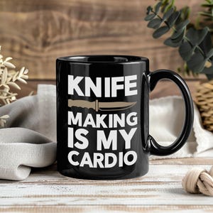 May include: Black ceramic mug with the text "KNIFE MAKING IS MY CARDIO" in white, with a tan knife graphic. The mug has a black handle and is set against a wooden background.