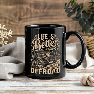 Op de afbeelding: Een zwarte keramische mok met een beige grafische print van een terreinwagen op een rotsachtig pad door een bos. De tekst "LIFE IS Better OFFROAD" is in een decoratief lettertype boven en onder de voertuigillustratie gedrukt.