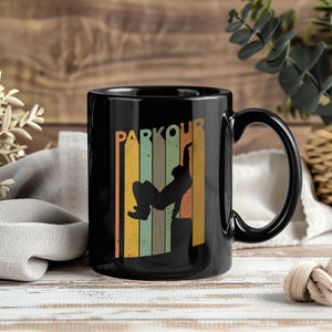 May include: Black coffee mug with a curved handle. The mug features the word "PARKOUR" in orange letters above a silhouette of a person performing parkour. The background has vertical stripes in shades of orange, green, and yellow.