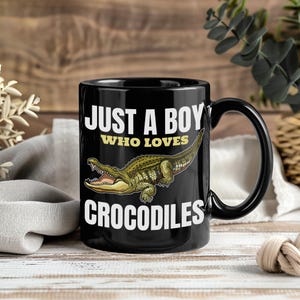 May include: Black ceramic mug with the text "JUST A BOY WHO LOVES CROCODILES" in white and yellow. A detailed illustration of a green and brown crocodile is featured on the mug.