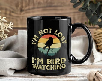 Puede incluir: Taza de cerámica negra con asa curva. La taza presenta el texto "I'M NOT LOST I'M BIRD WATCHING" en una fuente vintage, con una silueta de un pájaro posado en una rama dentro de un diseño de puesta de sol.