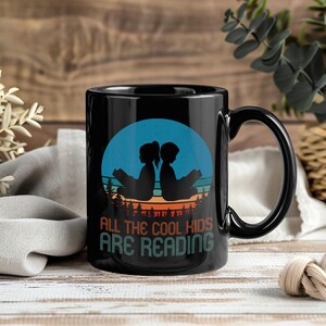 May include: Black ceramic mug with a wraparound graphic. The graphic features silhouettes of two children reading books, set against a blue and orange sunset. The text "ALL THE COOL KIDS ARE READING" is below the image.