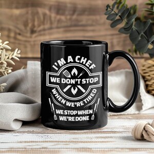 Peut inclure: Mug en céramique noire avec un graphisme blanc indiquant "I'M A CHEF WE DON'T STOP WHEN WE'RE TIRED WE STOP WHEN WE'RE DONE." La tasse a une grande anse et convient aux boissons chaudes ou froides. La tasse est sur une surface en bois.