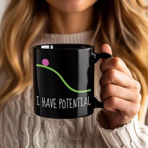 May include: Black ceramic mug with the phrase "I HAVE POTENTIAL" in white. The mug features a green line and a pink circle, resembling a physics diagram. The mug is held by a person.
