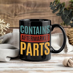 May include: Black ceramic mug with the text "CONTAINS AFTERMARKET PARTS" in teal, red, and yellow. The mug has a black handle and is set against a wooden background.